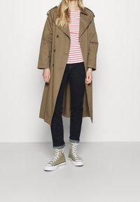 Abrigo trinchero caqui con grandes solapas, camisa a rayas rojas y blancas, jeans oscuros y zapatillas altas verdes con suelas y cordones blancos.