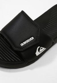 Čierne šľapky so štruktúrovanou podrážkou, syntetickým zvrškom a výrazným bielym logom "Quiksilver". Obsahujú zakrivený, nastaviteľný popruh.