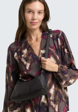 Schwarze Lederhandtasche mit einer einzigartigen Form, die eine strukturierte Oberfläche und einen dünnen, verstellbaren Tragegurt aufweist, gehalten von einem Model in einer gemusterten Bluse.