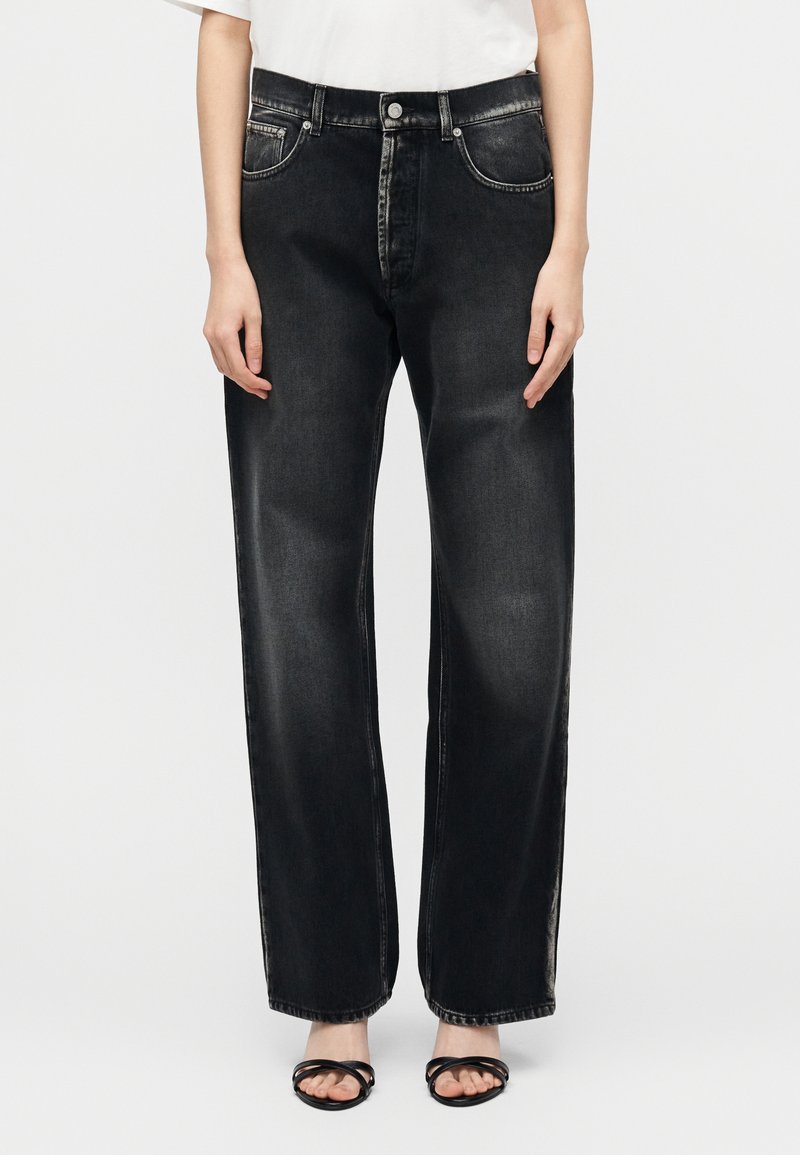 Άτομο που φοράει μαύρο ξεθωριασμένο τζιν με ψηλόμεση ίσια γραμμή και μαύρα σανδάλια ανοιχτά στο μπροστινό μέρος, στέκεται μπροστά σε απλό ανοιχτό υπόβαθρο.