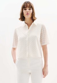 Korte mouw, gekraagde crèmekleurige blouse met een getextureerd zigzagpatroon. Voorzien van een knoopsluiting aan de voorkant en een relaxte pasvorm, gecombineerd met witte broeken.