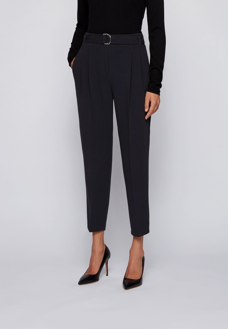 Nainen, joka on pukeutunut mustiin korkeavyötäröisiin räätälöityihin housuihin vyöllä, yhdistettynä mustaan pitkähiharaiseen paitaan ja mustiin korkokenkiin, seisoo yksinkertaisella taustalla.