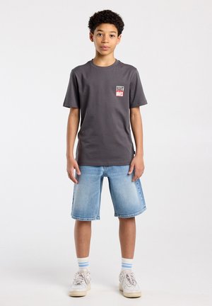 Jongen met krullend haar, een grijs T-shirt met print, knielange spijkershorts, gestreepte sokken en witte sneakers, staand tegen een effen achtergrond.