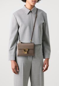 Brown leather crossbody bag with gold hardware, featuring a flap closure and a circular accent. Worn over a checkered patterned outfit.