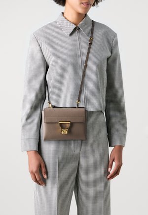 Smeđa kožna crossbody torba sa zlatnim detaljima, s preklopom i kružnim akcentom. Nosena preko odjeće s kariranim uzorkom.