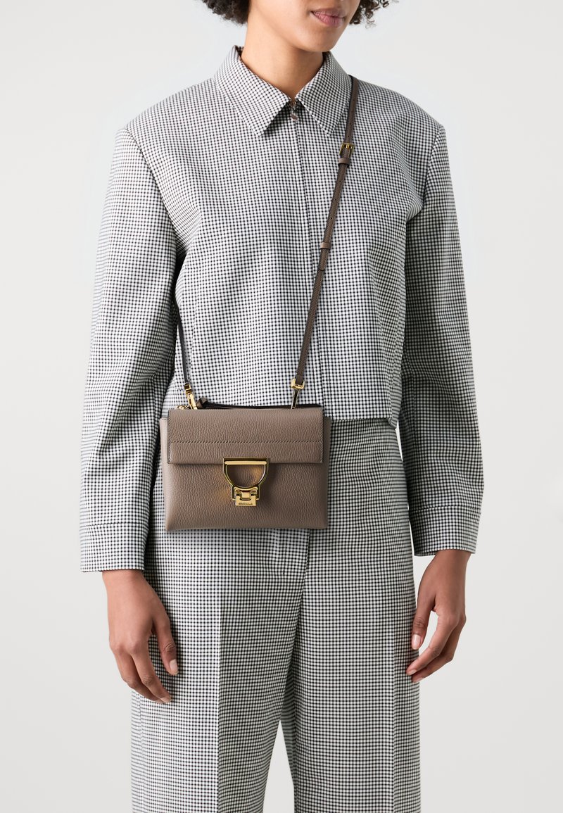 Brown leather crossbody bag with gold hardware, featuring a flap closure and a circular accent. Worn over a checkered patterned outfit.