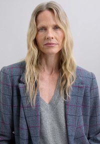 Blazer a cuadros grises en tejido texturizado con acentos en rosa, sobre un suéter gris de cuello en V. Cabello largo y ondulado en rubio enmarcando la cara.