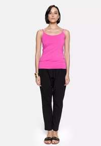 Różowa dopasowana podkoszulka na cienkich ramiączkach, zestawiona z luźnymi czarnymi spodniami. Modelka stoi w czarnych sandałach, z prostą złotą bransoletką.