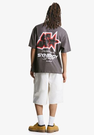 Grå grafisk t-shirt med rød stjernering, parret med hvide knæ-lange short. Modellen har brune sko og hvide sokker på, set fra bagfra.