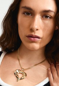 Collier avec pendentif en forme de cœur en or, au fini texturé, sur une chaîne épaisse, porté autour du cou sur un fond rayé noir et blanc.