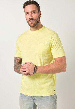 Uomo con barba e tatuaggi che indossa una maglietta gialla chiaro a maniche corte e jeans azzurri, in piedi davanti a uno sfondo semplice.