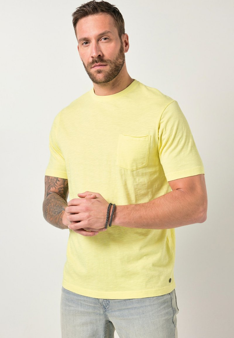 Man with beard and tattoos wearing a light yellow short-sleeve T-shirt and light blue jeans, standing against a plain background.