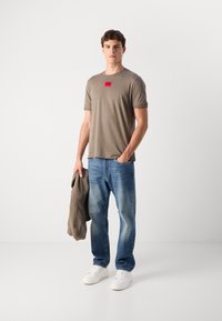 Tricou scurt cu mânecă maro, etichetă roșie, blugi albaștri decolorati și adidași albi; ținând un hanorac gri. Fundal simplu.