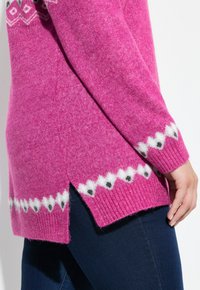 Różowy dziergany sweter z białym i czarnym wzorem rombów wzdłuż brzegu i mankietów, noszony z ciemnoniebieskimi dżinsami, ręka spoczywa wzdłuż boku.