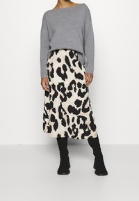 Nainen, joka on pukeutunut harmaaseen pitkähihainen yläosaan, beigen ja mustan eläinkuviolliseen midihameeseen sekä mustiin polven korkeisiin saappaisiin, seisoo valkoista taustaa vasten.