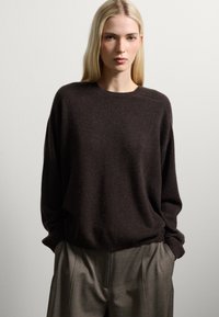 Maglione marrone scuro, con una trama morbida, colletto rotondo e spalle scese. Indossato sopra pantaloni grigi chiari e plissettati. Design semplice e pulito.