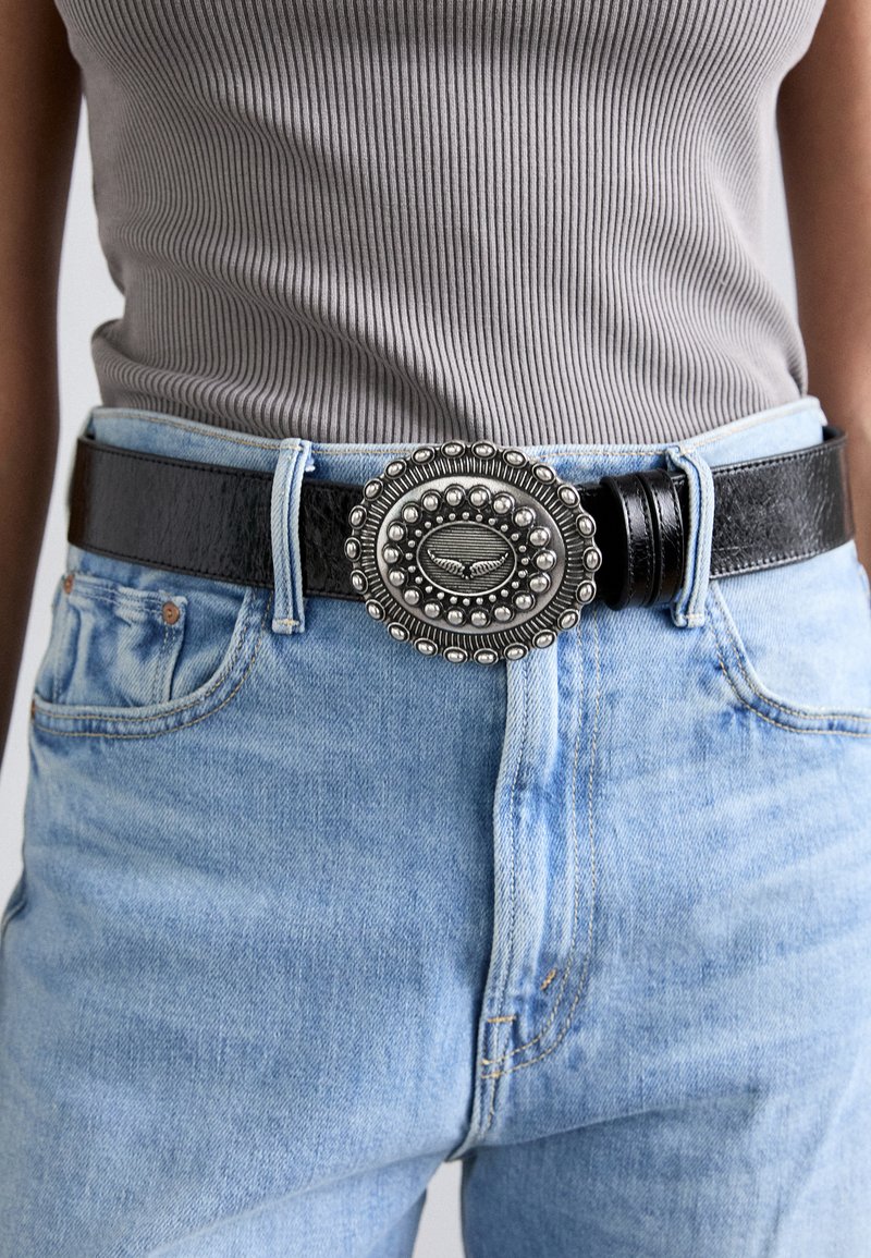 Black leather belt with a round, ornate silver buckle featuring raised details and beading, worn over light blue denim jeans.