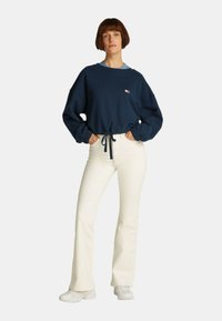 Felpa blu navy con polsini elasticizzati e un laccio in vita, abbinata a pantaloni a zampa d'elefante color crema e sneakers bianche. Outfit semplice e informale.