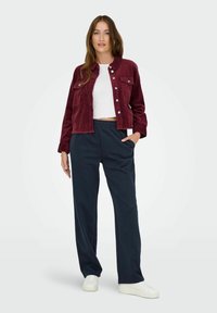 Bordeaux corduroy jasje met voorkanten, gecombineerd met marineblauwe relaxte broek. Model draagt een witte crop top en witte sneakers.