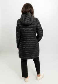 Cappotto imbottito nero con cappuccio, caratterizzato da cuciture verticali trapuntate e design di lunghezza media. Indossato sopra pantaloni neri e scarpe casual.