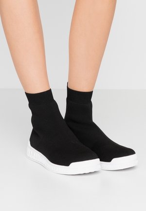 Benen met zwarte sneakers in sokstijl van gebreide stof met geribde manchetten en dikke witte zolen waarop merktekst is gegraveerd, tegen een effen grijze achtergrond.