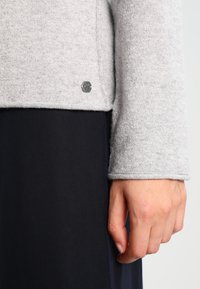 Jasnoszary sweter o miękkiej fakturze, z okrągłym metalowym logo na dole. Długi rękaw, noszony na ciemnogranatowym ubraniu.
