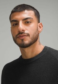 Schwarzer strukturierter Pullover mit Rundhalsausschnitt, getragen von einem Mann mit kurzen dunklen Haaren und einem Bart, vor einem neutralen grauen Hintergrund.