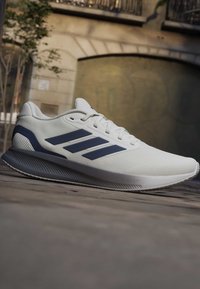 Weißer Laufschuh mit dunkelblauen Streifen und Sohle, mit der Aufschrift „Cloudfoam“, liegt auf einer steinernen Außenfläche in der Nähe eines Gebäudes.