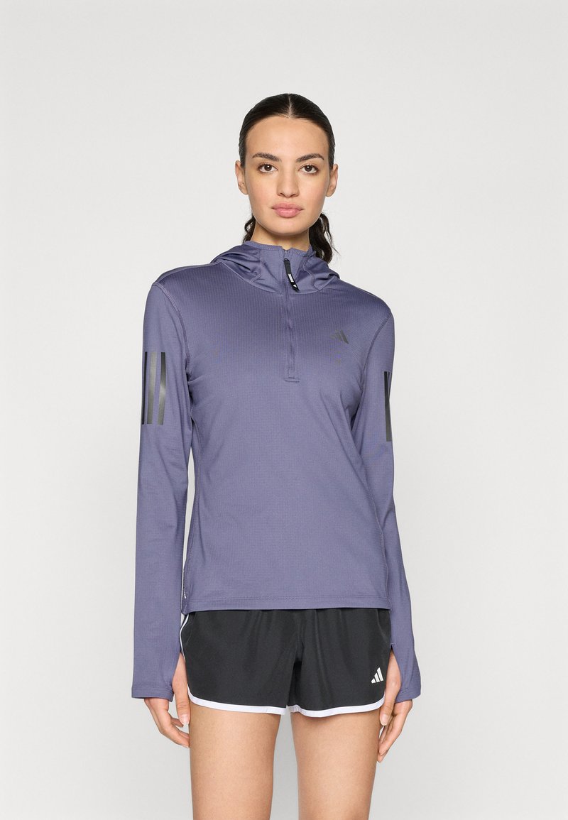 Haut de sport à manches longues violet avec capuche, fermeture éclair à mi-hauteur à l'avant, tissu texturé et accents noirs à trois rayures sur les manches. Porté avec un short noir.