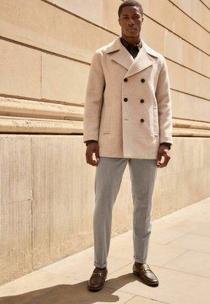 Lys beige dobbeltradet frakke med sorte knapper, grå skræddersyede bukser og brune loafers med hardware-detaljer. Glat tekstur og struktureret design.