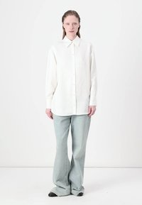 Camicia bianca a maniche lunghe con colletto a punta, bottoni frontali e vestibilità rilassata. Abbinata a jeans svasati azzurri e scarpe nere.