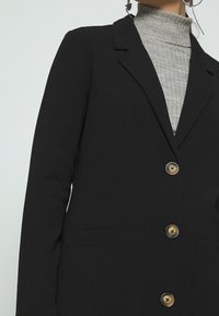 Černý blazer s texturovanou tkaninou, má klopový límec, tři knoflíky se zlatými akcenty a velké kapsy. Nositý přes šedý rolák.