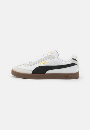 Baskets basses blanches Puma avec bande noire, semelle en gomme, lacets blancs et logos Puma dorés sur le côté, la languette et le talon.