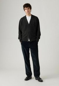 Dunkelgraue gestrickte Strickjacke mit V-Ausschnitt, zwei Knöpfen und gerippten Bündchen, kombiniert mit dunklen karierte Hosen und schwarzen Schuhen.