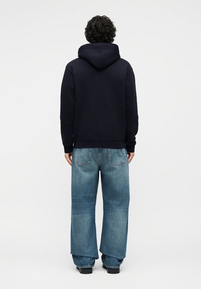 Personne aux cheveux bouclés portant un sweat à capuche noir et un jean bleu ample, debout de dos sur un fond uni.