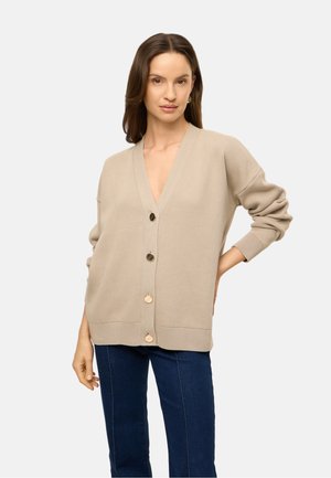 Vrouw met bruin haar die een beige oversized cardigan met vier knopen draagt en donkerblauwe spijkerbroek tegen een effen lichte achtergrond.