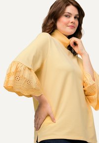 Top giallo con alta scollatura e maniche svasate, decorato con bordi in pizzo ad occhio. Tessuto morbido con una vestibilità comoda, spacco laterale sull'orlo.
