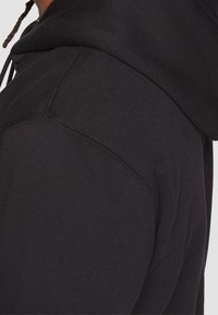 Svart huvtröja med en mjuk, texturerad tyg. Anmärkningsvärda detaljer inkluderar en justerbar huva och förstärkta sömmar vid axlarna.