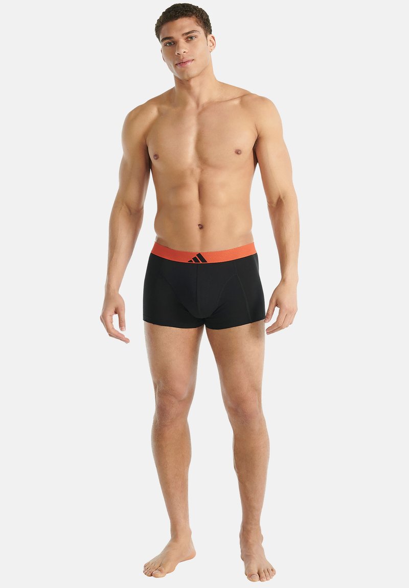 Homme torse nu portant un boxer noir avec une ceinture orange et un logo, debout pieds nus contre un fond blanc.