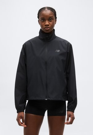 Žena s pletenými vlasmi, oblečená v čiernej bunde na zips a čiernych šortkách, stojaca na jednoduchom svetlom pozadí.