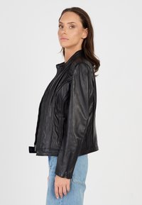 Zwarte leren motorjack met een ritssluiting aan de voorkant, een aansluitend ontwerp en ritssluitingen bij de manchetten. Draagt met lichtblauwe jeans, wat een slanke silhouet toont.