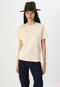 Beige bomull T-skjorte med rund hals, korte ermer og løs passform, kombinert med mørk denim jeans og en olivengrønn filthat.