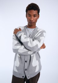Gray cardigan with white zigzag pattern, ribbed cuffs and hem, and black buttons. Worn over a gray shirt, featuring a layered collar.