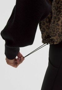 Sort ribbet sweater med voluminøse ærmer, parret med en brun leopardmønstret jakke. En snor bliver trukket i jackens bund.