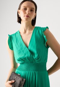 Green dress with a deep V-neck, ruffled sleeves, and smocked waist. Model holds a silver fringe clutch. Smooth fabric and flowing design.