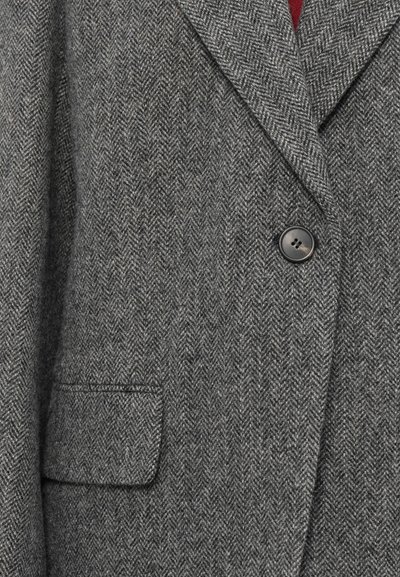 Grå herringbone-blazer i ull, med två knappar, slagkrage och en enkel bröstficka. Den texturerade tyget ger djup.