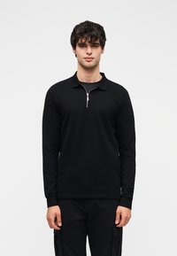 Μαύρο πόλο με μακριά μανίκια και φερμουάρ στο γιακά. Το ύφασμα φαίνεται λείο και το σχέδιο είναι απλό και εφαρμοστό. Φοριέται από ένα μοντέλο που στέκεται όρθιο.