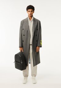 Grauer, doppelt-knöpfender Mantel mit schwarzen Knöpfen, getragen über einer hellgrauen Zip-Jacke und beigen Jogginghosen. Schwarzer strukturierten Rucksack, weiße Turnschuhe.