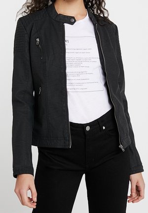 Femme portant une veste noire zippée sur un t-shirt blanc imprimé et un pantalon noir, debout les bras détendus le long du corps.