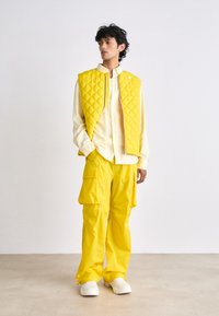 Gilet matelassé jaune associé à une chemise boutonnée jaune clair et un pantalon cargo assorti. Il comprend une fermeture éclair et des poches spacieuses.
